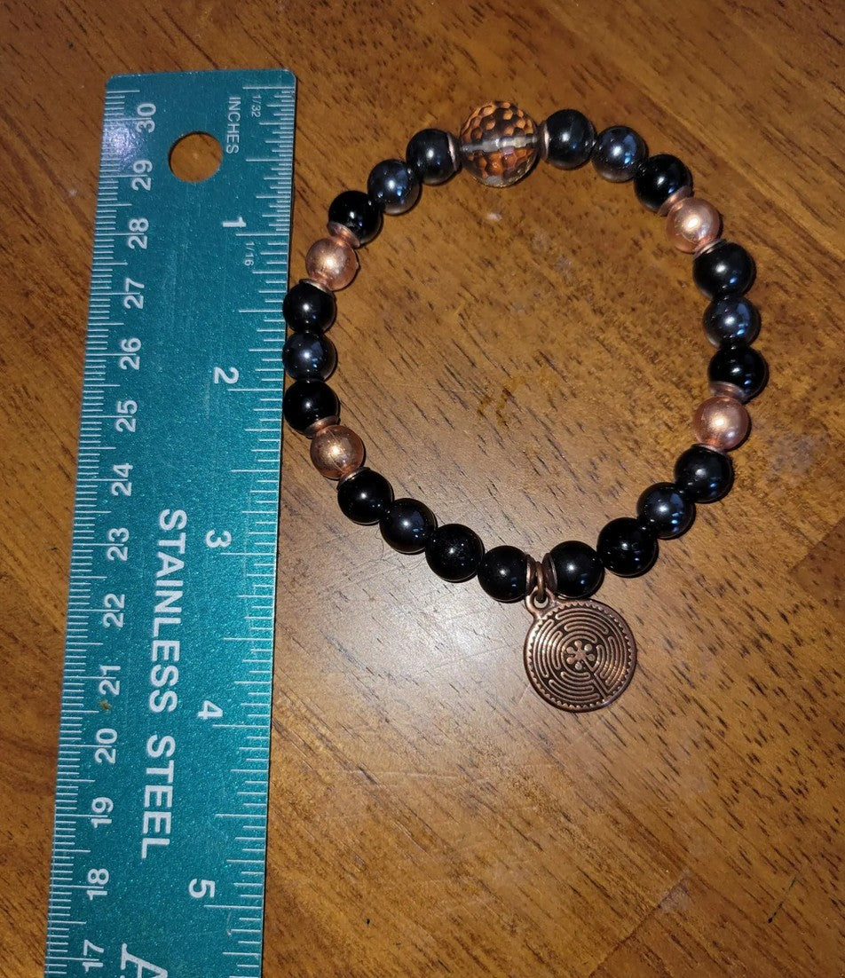 Black and copper beaded spiritual bracelet with charm beside a stainless steel ruler on wood table