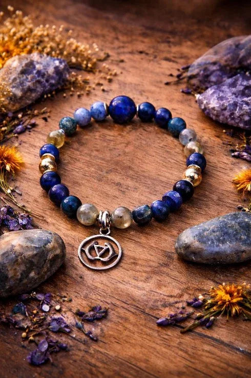 Beaded gemstone bracelet with silver charm on wooden surface, surrounded by crystals and flowers.