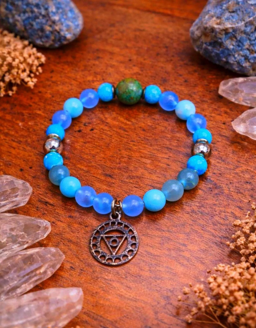 Blue chakra bead bracelet with silver charm on wooden surface, surrounded by crystals and stones