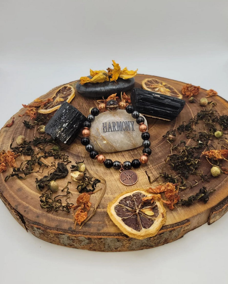 Spiritual bracelet with black and copper beads on wood slab, surrounded by crystals, dried herbs, and dried citrus slices for wellness and harmony.