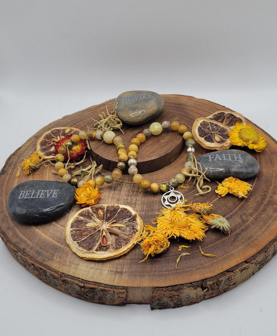 Yellow gemstone spiritual bracelet with silver charm on wooden display, surrounded by dried flowers, sliced citrus, and engraved stones reading believe, inspire, faith.