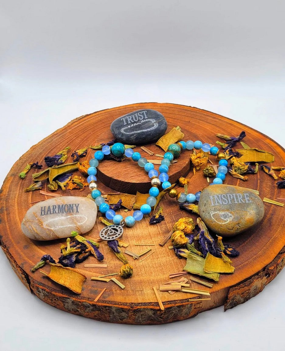 Blue spiritual bracelet with charm on wooden slab, surrounded by stones labeled Trust, Harmony, Inspire and dried flowers